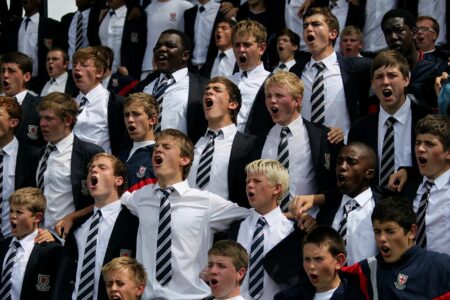 A photo of a group of school boys, all dressed in white shirts with black and silver striped ties. They all are mid-shouting.